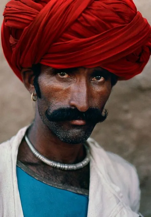 A man in a deep red turban wound high and full, looking straight out. A heavy moustache, a gold ring in one ear, a silver torque at his throat, a white cloth thrown over a blue shirt.