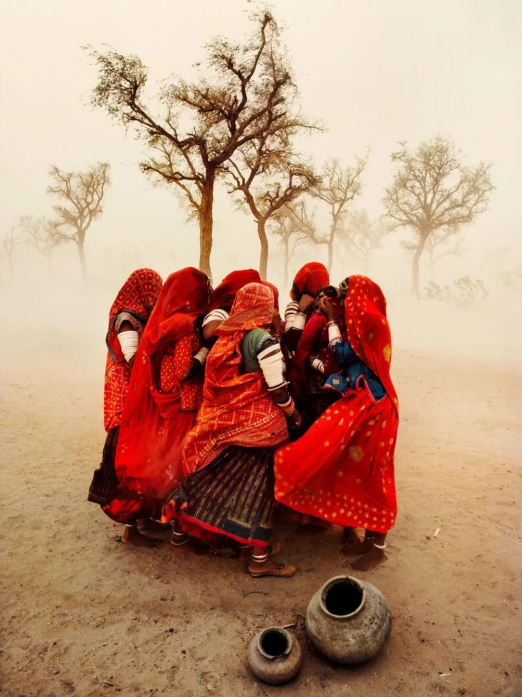 A knot of women in red veils crouched together in the open, holding on to one another as a dust storm blows through. Thorn trees bend behind them in the yellow air; two clay water pots stand on the ground in front.